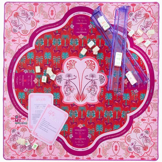 A colorful board game set laid out flat on a white background. The square game board has a pink and red patterned design with floral motifs and several translucent purple dice trays containing white dice.